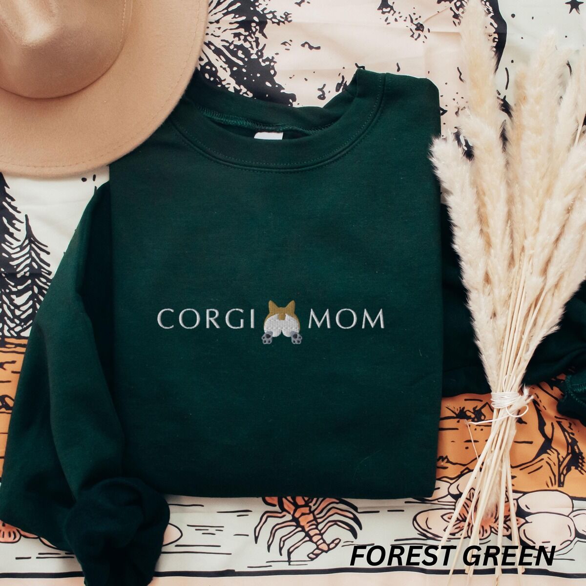 Corgi Butt Sweatshirt – Forest Green Forest Green crewneck sweatshirt laid flat with a felt hat and dried pampas grass, embroidered with CORGI MOM in light thread around a corgi butt
