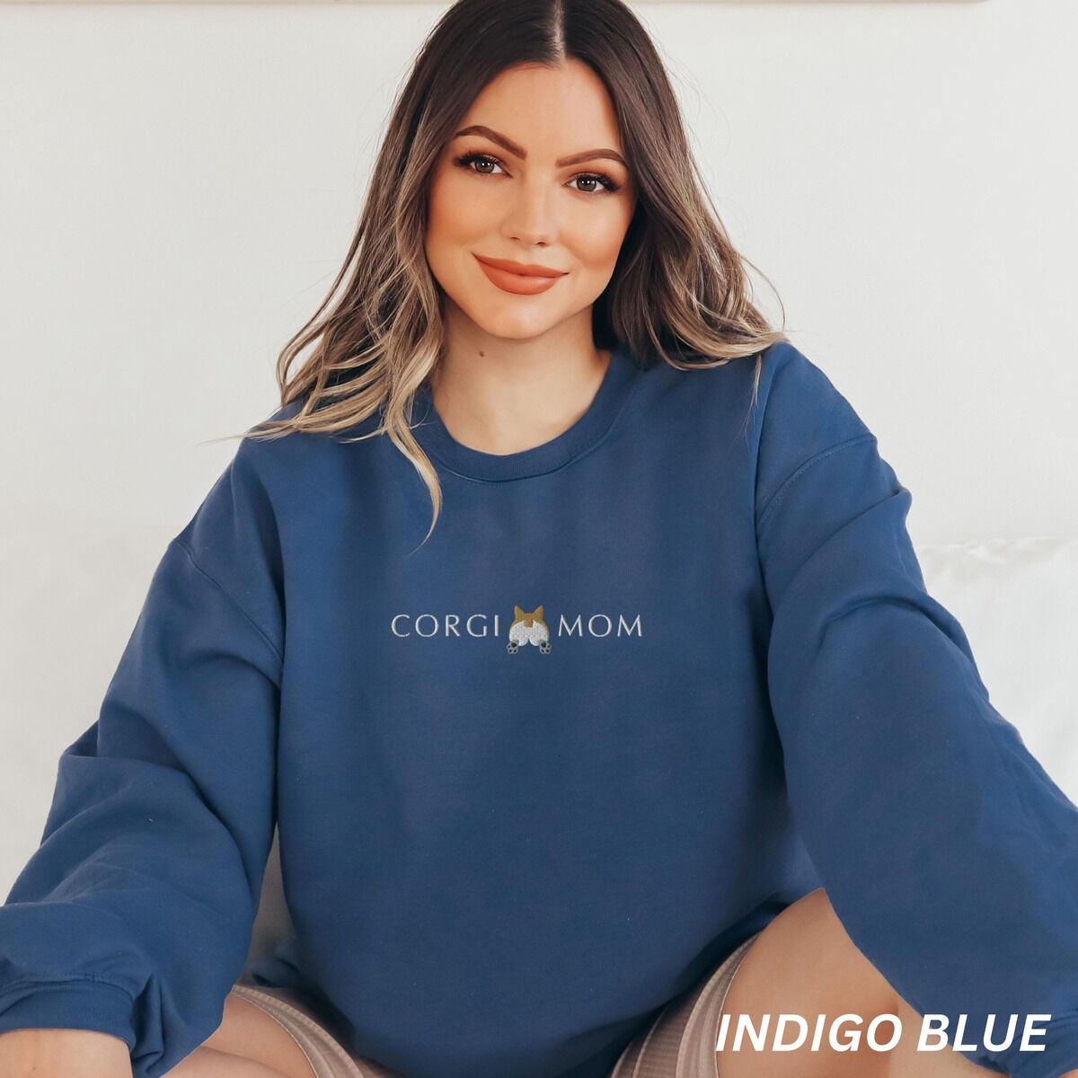 Corgi Butt Sweatshirt – Indigo Blue Smiling woman in an Indigo Blue crewneck sweatshirt embroidered with CORGI MOM and a corgi butt between the words