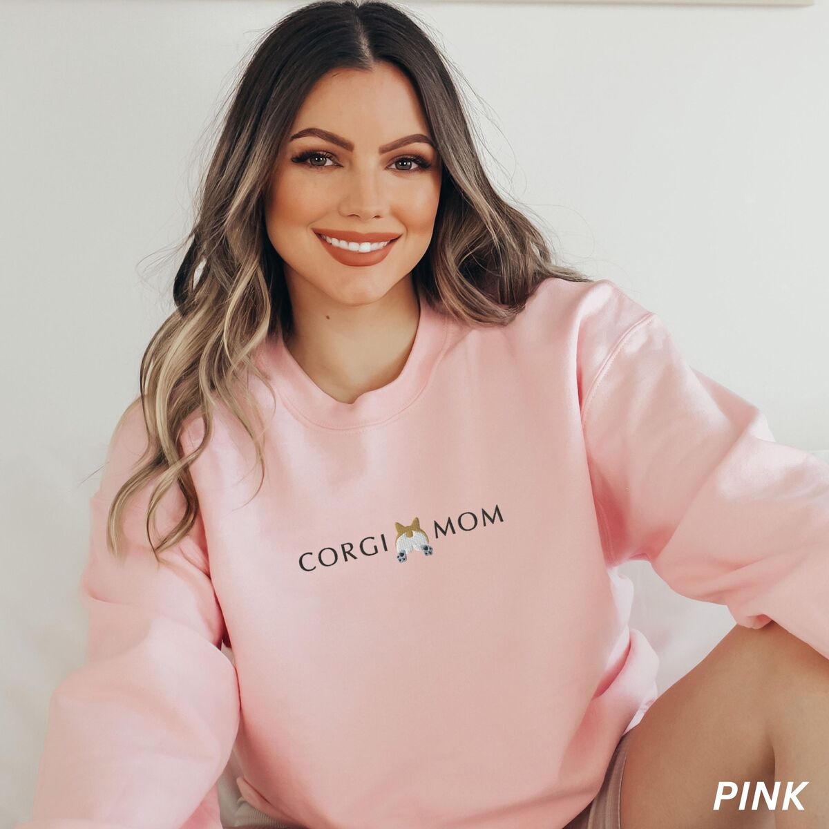 Corgi Butt Sweatshirt - Light Pink Smiling woman in a Light Pink crewneck sweatshirt embroidered with CORGI MOM and a corgi butt with two little paws