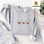 Ash crewneck sweatshirt folded with denim shorts, white sneakers and a potted plant, embroidered with three tricolor corgis in red knit scarves among sprigs of holly