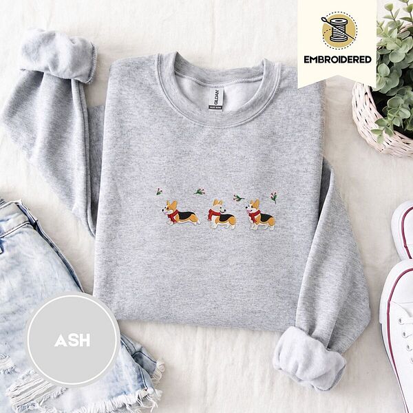 Ash crewneck sweatshirt folded with denim shorts, white sneakers and a potted plant, embroidered with three tricolor corgis in red knit scarves among sprigs of holly