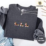 Dark Heather crewneck sweatshirt folded with ripped jeans and a silver chain necklace, embroidered with three tricolor corgis in red knit scarves among sprigs of holly