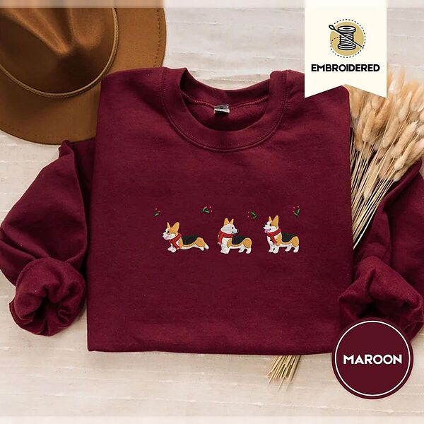 Maroon crewneck sweatshirt with a brown felt hat and dried bunny tail grass, embroidered with three tricolor corgis in red knit scarves among sprigs of holly