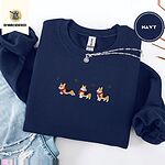 Navy crewneck sweatshirt folded with ripped jeans and a silver chain necklace, embroidered with three tricolor corgis in red knit scarves among sprigs of holly