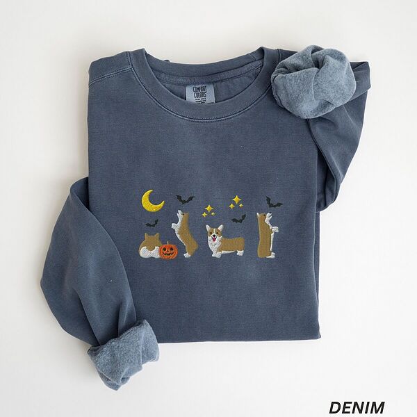 Denim crewneck sweatshirt laid flat with a rolled sleeve, embroidered with four corgis under a crescent moon with bats, gold sparkles and a jack-o'-lantern