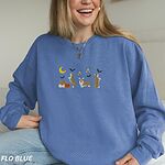 Smiling woman sitting in a Flo Blue crewneck sweatshirt, embroidered with four corgis under a crescent moon with bats, gold sparkles and a jack-o'-lantern