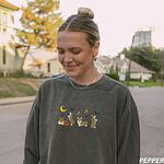 Woman wearing a Pepper crewneck sweatshirt on a tree-lined street, embroidered with four corgis under a crescent moon with bats, gold sparkles and a jack-o'-lantern
