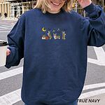 Smiling woman holding a coffee in a True Navy crewneck sweatshirt on a city street, embroidered with four corgis under a crescent moon with bats, gold sparkles and a jack-o'-lantern