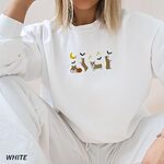 Woman sitting in a White crewneck sweatshirt, embroidered with four corgis under a crescent moon with bats, gold sparkles and a jack-o'-lantern