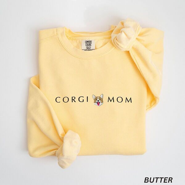 Butter crewneck sweatshirt laid flat on white, embroidered with CORGI MOM in dark thread and a red and white corgi face with its tongue out