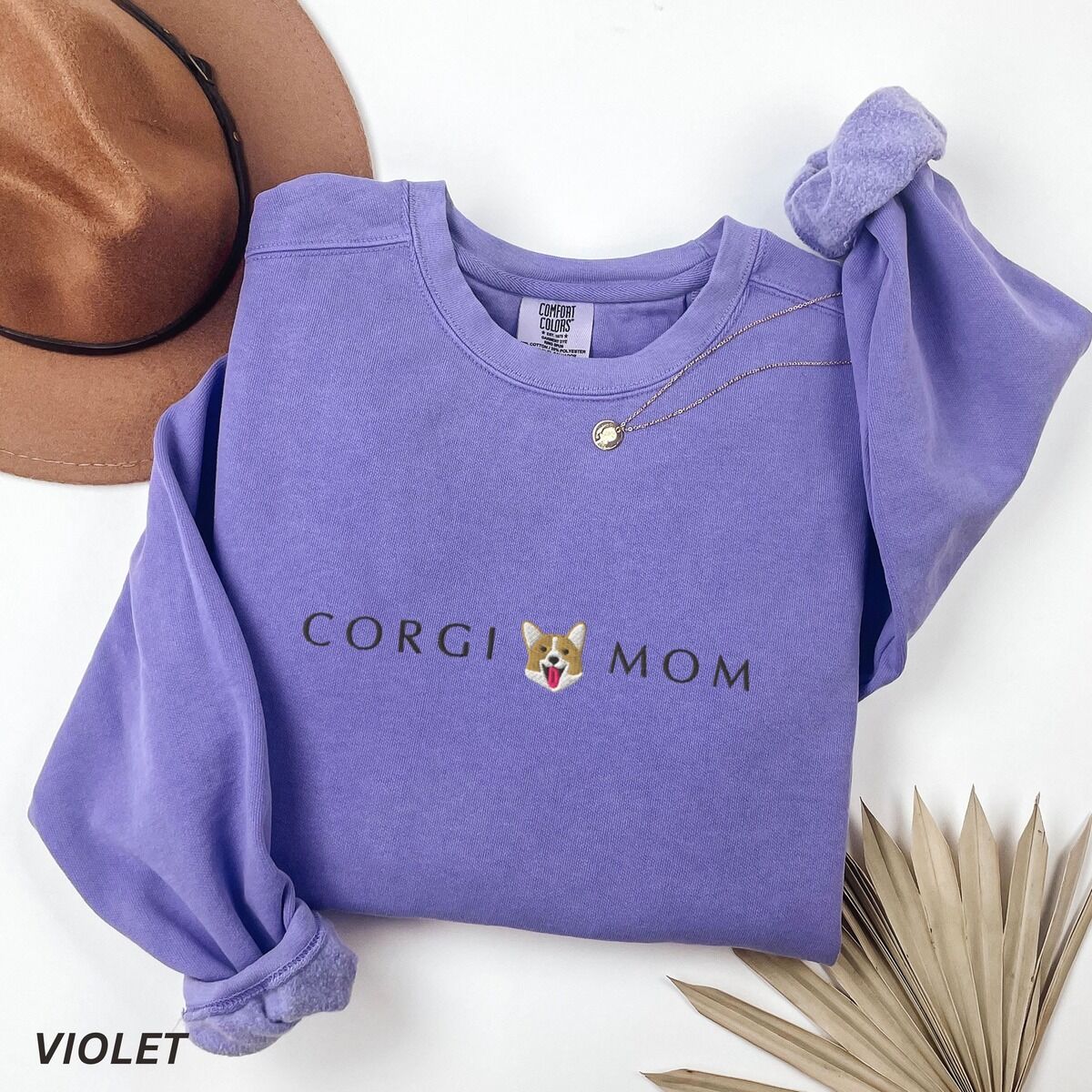 Corgi Mom Sweatshirt – Violet Violet crewneck sweatshirt laid flat with a felt hat and dried palm leaves, embroidered with CORGI MOM and a smiling corgi face