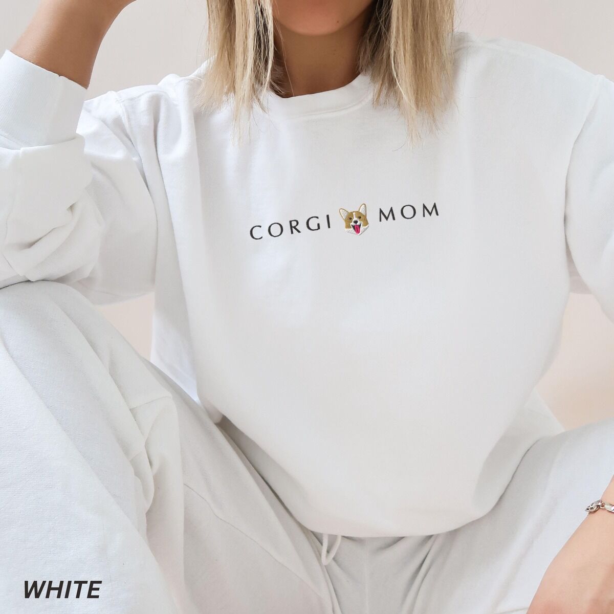 Corgi Mom Sweatshirt – White Woman sitting in a White crewneck sweatshirt embroidered with CORGI MOM and a smiling corgi face across the chest