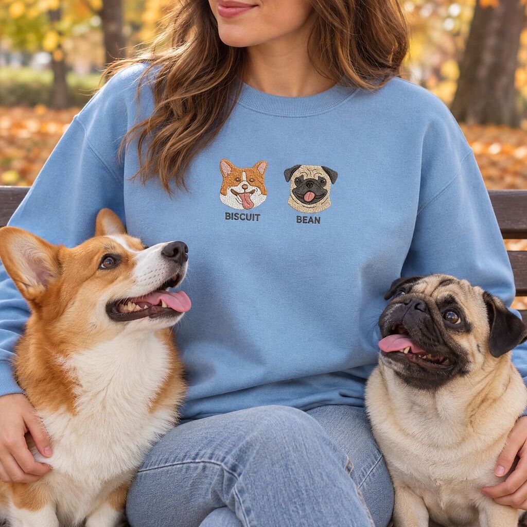 Woman on a park bench in a Light Blue sweatshirt embroidered with a corgi and a pug, sitting with her own corgi and pug