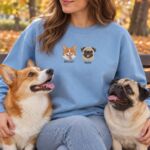 Woman on a park bench in a Light Blue sweatshirt embroidered with a corgi and a pug, sitting with her own corgi and pug