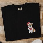Black T-shirt laid flat on wood beside white sneakers, embroidered with a smiling red and white Pembroke corgi in a Santa hat and striped red scarf on the left chest