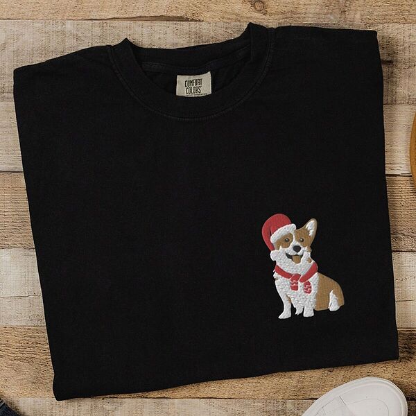 Black T-shirt laid flat on wood beside white sneakers, embroidered with a smiling red and white Pembroke corgi in a Santa hat and striped red scarf on the left chest