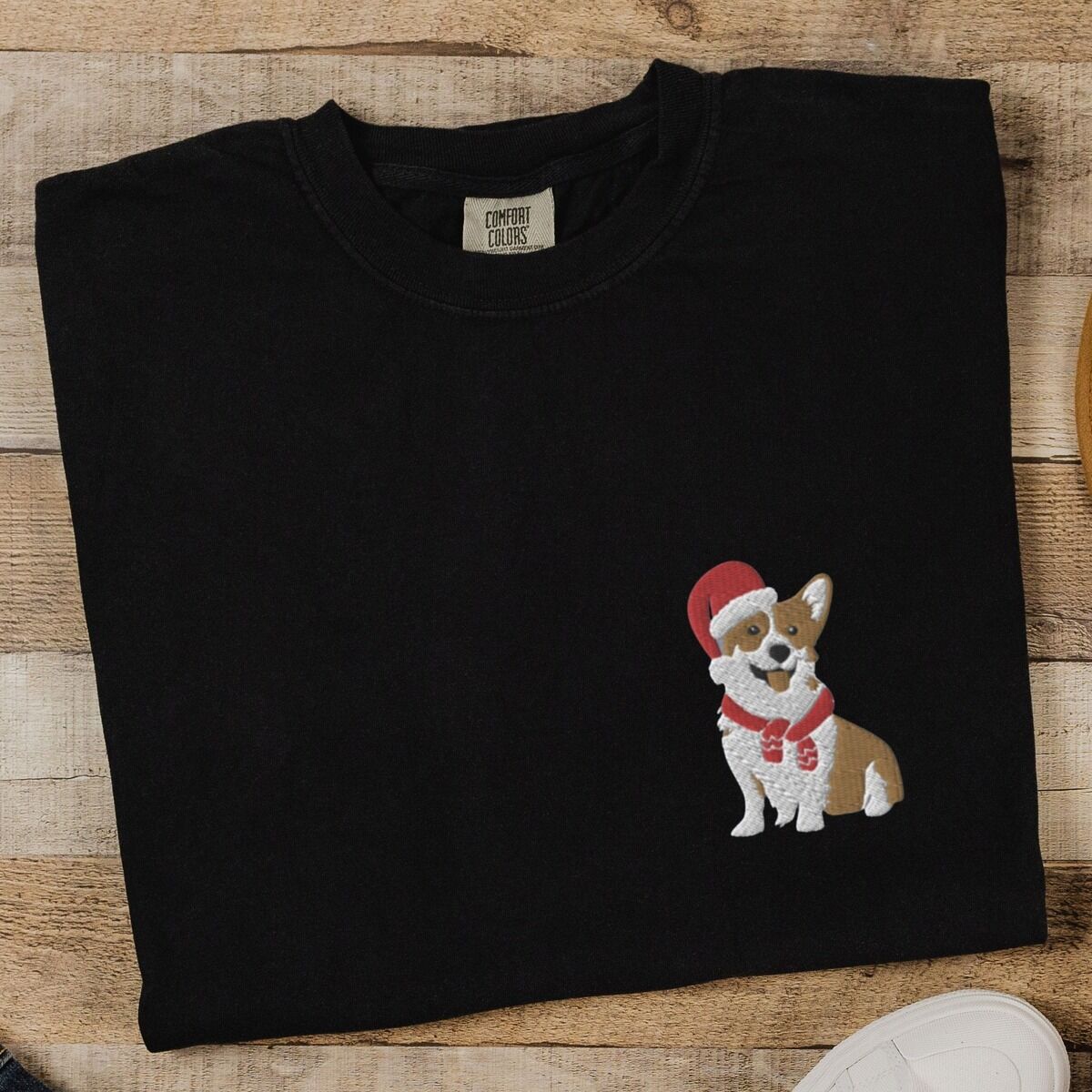 Corgi Santa Hat Shirt – Black Black T-shirt laid flat on wood beside white sneakers, embroidered with a smiling red and white Pembroke corgi in a Santa hat and striped red scarf on the left chest
