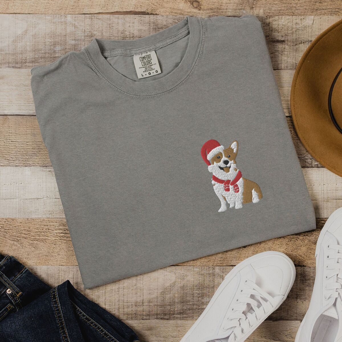 Corgi Santa Hat Shirt - Grey Grey T-shirt laid flat on wood beside jeans, a felt hat and white sneakers, embroidered with a smiling red and white Pembroke corgi in a Santa hat and striped red scarf on the left chest