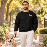 Man in a black embroidered corgi sweatshirt walking a red and white Pembroke Welsh corgi on a leash