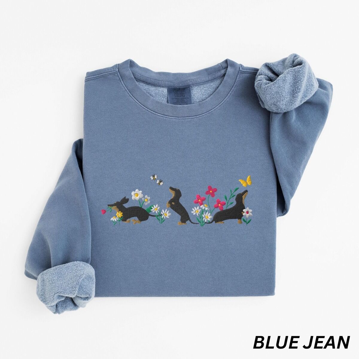 Cottagecore Dachshund Sweatshirt – Blue Jean Blue Jean crewneck sweatshirt laid flat on white, embroidered with three black and tan dachshunds playing among white daisies and pink spring flowers, with two bees and a yellow butterfly, across the chest