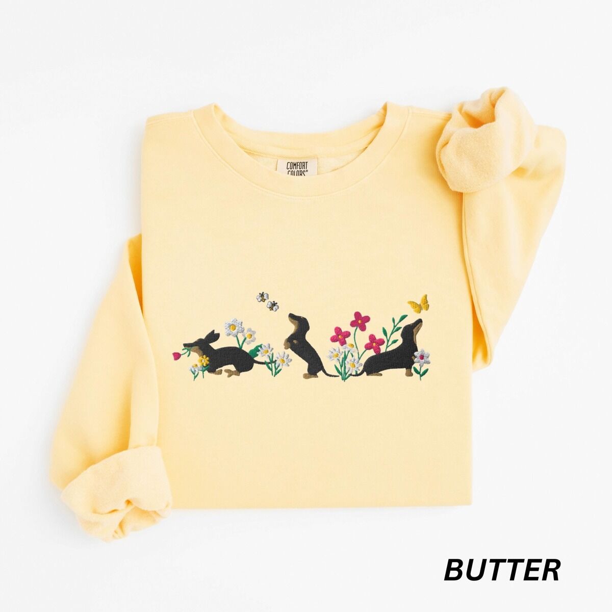 Cottagecore Dachshund Sweatshirt – Butter Butter crewneck sweatshirt laid flat on white, embroidered with three black and tan dachshunds playing among white daisies and pink spring flowers, with two bees and a yellow butterfly, across the chest