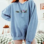 Woman in an oversized Blue Jean crewneck sweatshirt embroidered with three black and tan dachshunds playing among white daisies and pink spring flowers, with two bees and a yellow butterfly, across the chest