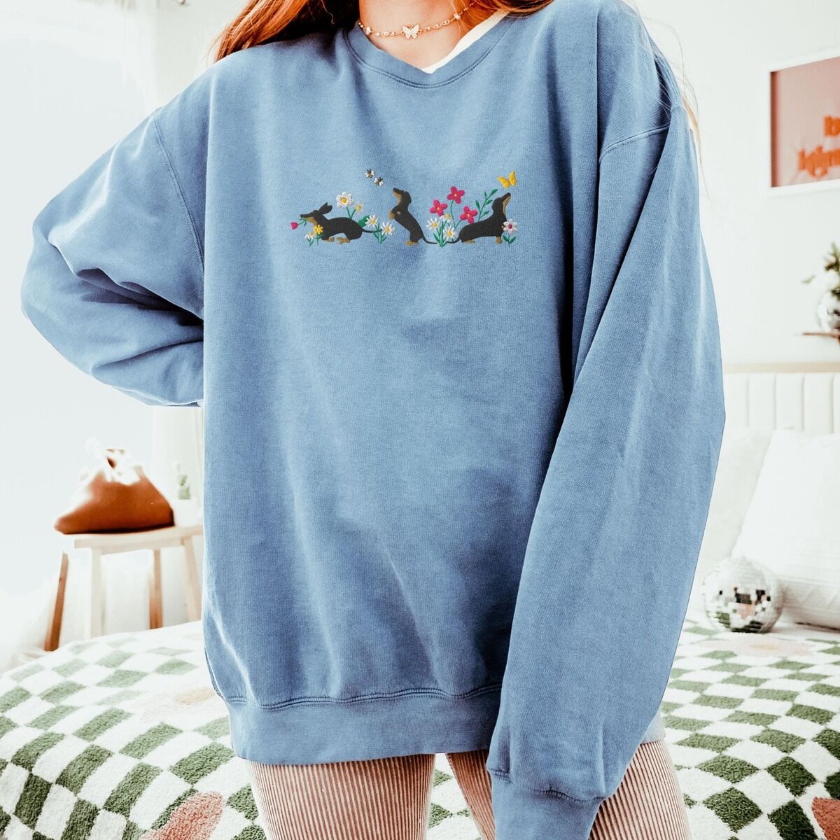 Cottagecore Dachshund Sweatshirt - worn in blue jean Woman in an oversized Blue Jean crewneck sweatshirt embroidered with three black and tan dachshunds playing among white daisies and pink spring flowers, with two bees and a yellow butterfly, across the chest