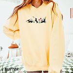 Woman in an oversized Butter crewneck sweatshirt embroidered with three black and tan dachshunds playing among white daisies and pink spring flowers, with two bees and a yellow butterfly, across the chest