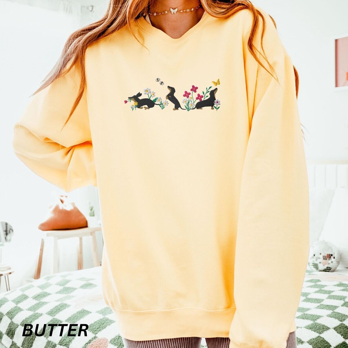 Cottagecore Dachshund Sweatshirt - worn in butter Woman in an oversized Butter crewneck sweatshirt embroidered with three black and tan dachshunds playing among white daisies and pink spring flowers, with two bees and a yellow butterfly, across the chest