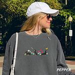 Woman in a cap on a city street wearing a Pepper crewneck sweatshirt embroidered with three black and tan dachshunds playing among white daisies and pink spring flowers, with two bees and a yellow butterfly, across the chest