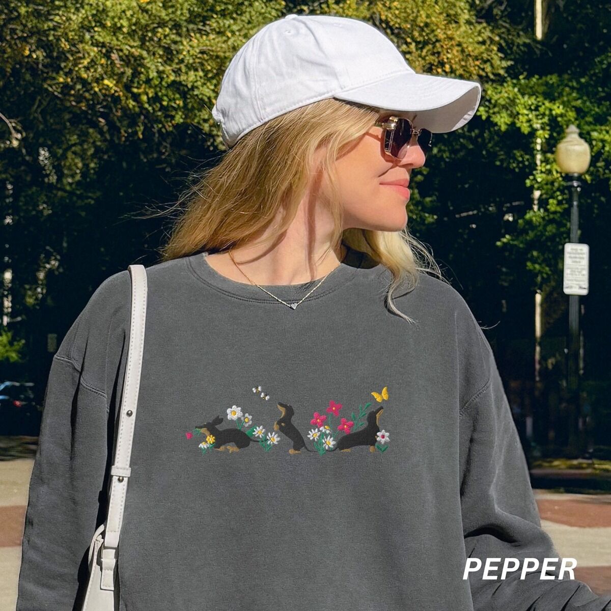 Cottagecore Dachshund Sweatshirt - worn in pepper Woman in a cap on a city street wearing a Pepper crewneck sweatshirt embroidered with three black and tan dachshunds playing among white daisies and pink spring flowers, with two bees and a yellow butterfly, across the chest