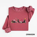 Crimson crewneck sweatshirt laid flat on white, embroidered with three black and tan dachshunds playing among white daisies and pink spring flowers, with two bees and a yellow butterfly, across the chest