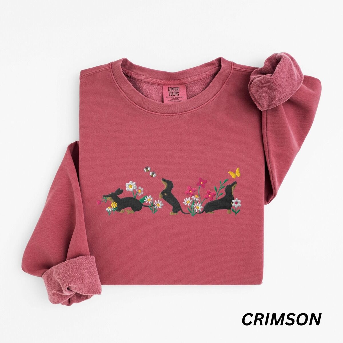 Cottagecore Dachshund Sweatshirt – Crimson Crimson crewneck sweatshirt laid flat on white, embroidered with three black and tan dachshunds playing among white daisies and pink spring flowers, with two bees and a yellow butterfly, across the chest