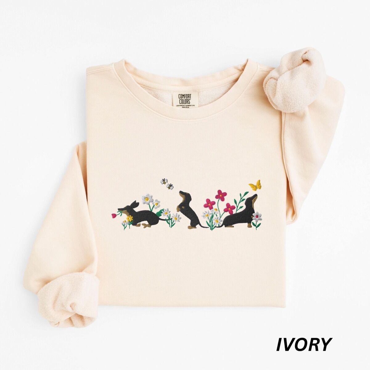 Cottagecore Dachshund Sweatshirt – Ivory Ivory crewneck sweatshirt laid flat on white, embroidered with three black and tan dachshunds playing among white daisies and pink spring flowers, with two bees and a yellow butterfly, across the chest
