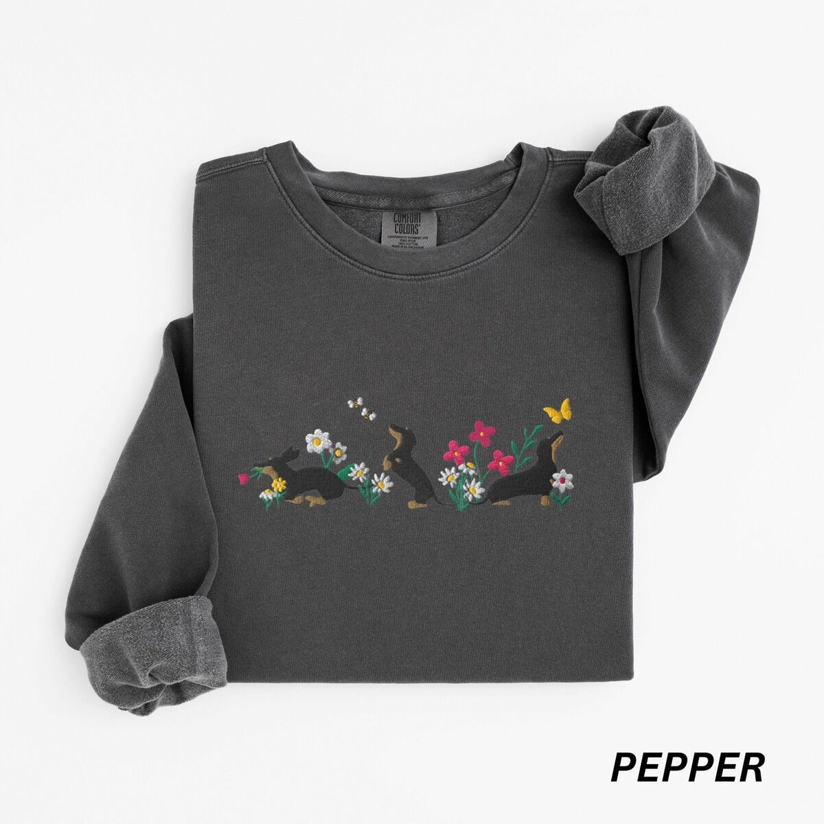 Cottagecore Dachshund Sweatshirt – Pepper Pepper crewneck sweatshirt laid flat on white, embroidered with three black and tan dachshunds playing among white daisies and pink spring flowers, with two bees and a yellow butterfly, across the chest