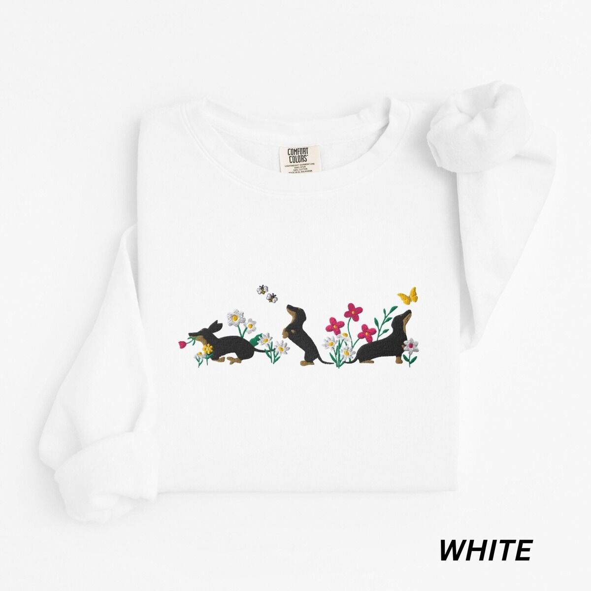 Cottagecore Dachshund Sweatshirt – White White crewneck sweatshirt laid flat on white, embroidered with three black and tan dachshunds playing among white daisies and pink spring flowers, with two bees and a yellow butterfly, across the chest