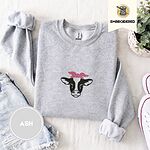 Ash crewneck sweatshirt folded with denim shorts, white sneakers and a potted plant, embroidered with a black and white cow head wearing a pink heart-print bandana tied in a bow