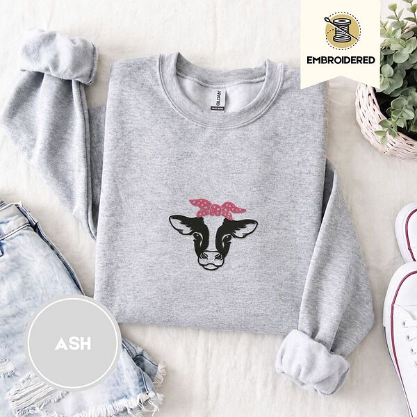 Ash crewneck sweatshirt folded with denim shorts, white sneakers and a potted plant, embroidered with a black and white cow head wearing a pink heart-print bandana tied in a bow
