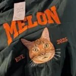 Green crewneck with an embroidered orange cat face, the name MELON in orange varsity letters and EST 2025