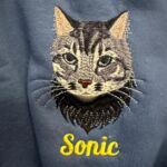 Close-up of an embroidered tabby cat portrait with the name Sonic in yellow script on a blue sweatshirt