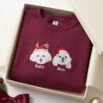 Maroon custom dog Christmas sweatshirt with two embroidered white dog portraits in a Santa hat and reindeer antlers, named Ruby and Max, in a gift box