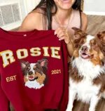 Woman holding a maroon sweatshirt embroidered with her Australian shepherd Rosie, sitting next to the real dog