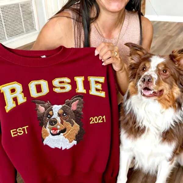 Woman holding a maroon sweatshirt embroidered with her Australian shepherd Rosie, sitting next to the real dog