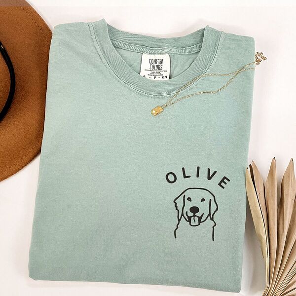Bay T-shirt laid flat with a necklace and a brown hat, embroidered with a line art golden retriever face and the name OLIVE on the left chest