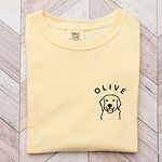 Butter T-shirt laid flat on light wood, embroidered with a line art golden retriever face and the name OLIVE on the left chest
