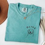Seafoam T-shirt laid flat with a necklace and a brown hat, embroidered with a line art golden retriever face and the name OLIVE on the left chest