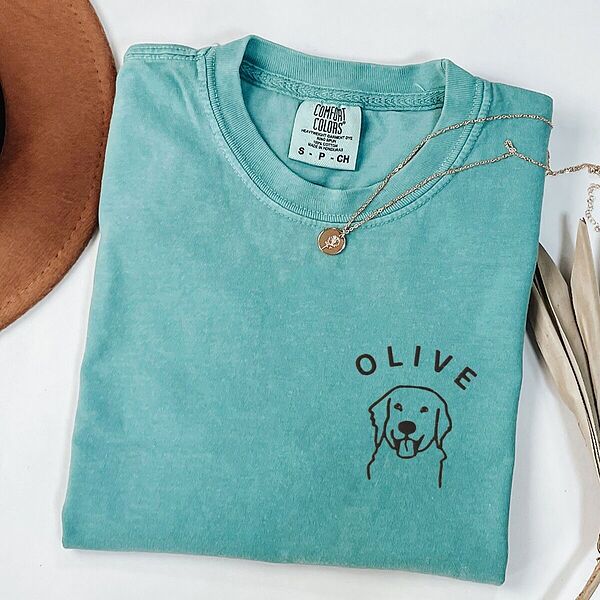 Seafoam T-shirt laid flat with a necklace and a brown hat, embroidered with a line art golden retriever face and the name OLIVE on the left chest