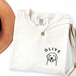 White T-shirt laid flat with a necklace and a brown hat, embroidered with a line art golden retriever face and the name OLIVE on the left chest