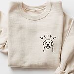 Sand crewneck sweatshirt laid flat with a rolled cuff, embroidered with a line art golden retriever face and the name OLIVE on the left chest