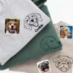 Three folded tops with line art pet portraits: a beagle named Cookie on grey, a golden retriever on green and a pug on sand